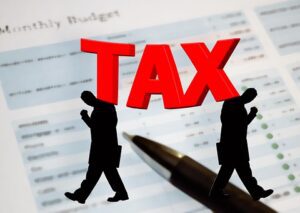Two silhouettes carry a large red TAX over a budget document and pen.