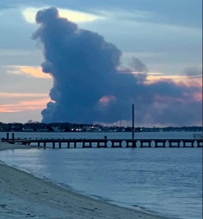 Large plume of dark smoke rising near a coastal area at sunset.