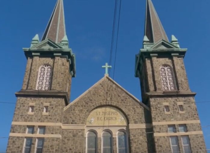Front view of a stone church with twin spires and a cross.
